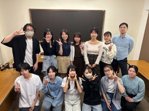 萩野谷ゼミ中業写真。教室で黒板を背景に、12人のゼミ生と萩野谷先生が2列に並びピースなどをしている