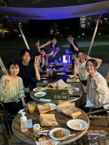 9人のゼミ生が夜に屋外で撮った写真。ジュースやカクテルのような色の飲み物や焼きそばなどが机上に広げられている。