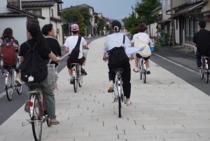 佐藤ゼミ、合宿の様子2。民家に囲まれた道路を自転車で走行するゼミ生たちの後ろ姿。