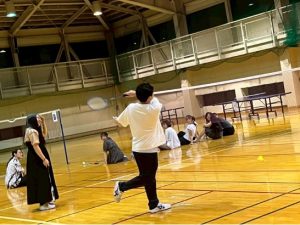 体育館でバドミントンをしている男性の後ろ姿の写真。周囲で7名が振り向き男性の方を見ている。