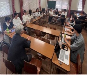 教室の中で、野末先生と約10名の学生が、コの字型に配置された長机を囲んで座っている様子。学生の多くはノートパソコンを開き、真剣な面持ちで教員や他の学生の話を聞いている。野末先生は手前の中央に座り、両手を組んで学生たちに向き合っている。