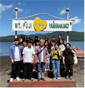 富士山の麓にある山中湖畔で、ゼミの学生と教員と思われる約20名のグループが、記念撮影用の看板の前に並んで立っている写真。看板には「MT. FUJI 山中湖 YAMANAKAKO」と書かれ、中央には黄色いハートのマークがある。皆、楽しそうにカメラに向かってポーズを取っており、背景には穏やかな湖面と緑豊かな山々が広がる。