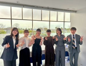 窓際の明るい場所で、フォーマルな服装またはビジネスカジュアルな服装をした女性5名と男性1名が並んで立っている写真。全員が笑顔でカメラに向かってピースサインをしている。