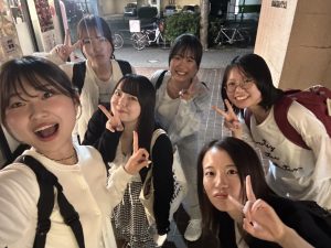 屋外で撮影された、6人の若い女性の集合写真（自撮り/セルフィー）。全員が笑顔でピースサインをしており、背景には店舗や自転車が写っている。
