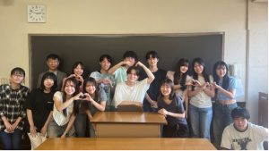宮本ゼミの集合写真。教室の黒板の前で撮影されており、宮本先生は後方左端に立っている。14名のゼミ生たちは笑顔で数名の学生は両手でハートマークを作るポーズをしており、和やかで親密な雰囲気が伝わる。
