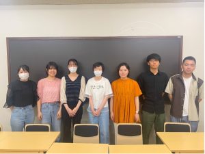 7名のゼミ生が、教室の黒板の前に一列に並んで立っている写真。手前には机と椅子が並んでいる。全員がカメラに向かって立っており、真面目だが少し微笑んでいる。