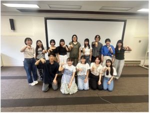 伊藤先生と13名が2列に並んで、片手の人差し指、中指、薬指を下向きに立てている。