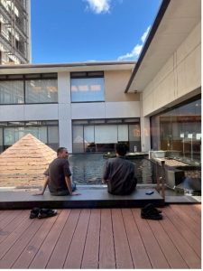 屋外にある建物のテラスで、男性2名が縁に座って水場を眺めている様子。現代的な建物と和風の庭園が組み合わされた空間で、リラックスしている雰囲気が伝わる。