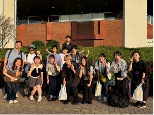 萩野谷ゼミの屋外での集合写真。煉瓦造りの建物と植え込みを背景に、萩野谷先生と13人のゼミ生たちが集まっている。ほとんどの学生がピースサインをしており、明るく楽しそうな雰囲気。