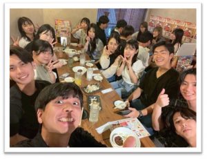 居酒屋のような場所でゼミ生たちがテーブルを囲み、食事を楽しんでいる自撮り写真。多くの学生がカメラに顔を向け、ピースサインや親指を立てるポーズ（いいね！）をして、楽しい雰囲気が伝わってくる。