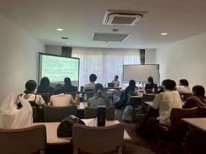 会議室で、大勢の参加者がスクリーンに映し出された資料を見ながら講義を受けている様子の写真。前には小林先生が座っている。