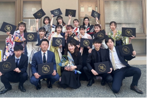 大学の建物の前で撮られた卒業式の集合写真。スーツや袴姿のゼミ生たちが、卒業証書（または証書ホルダー）を手に、笑顔でポーズを取っている。中央には花束を持った女性もいる。卒業証書ホルダーには**「MEIJI GAKUIN UNIVERSITY」**の文字が見える。