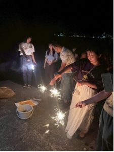 森本ゼミの学生たちが夜の屋外で手持ち花火を楽しんでいる様子。数名が明るい火花を上げる花火を手に持ち、笑顔でその光を見つめている。地面にはバケツなどの花火の道具が見える。