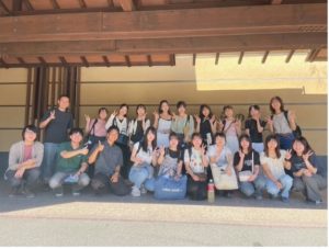 森本ゼミの大人数の学生と教員が、和風の建物の軒下と思われる場所で撮った集合写真。ほとんどの人がピースサインや親指を立てるポーズをして、明るい笑顔を見せている。ゼミ合宿の集合写真と思われる。