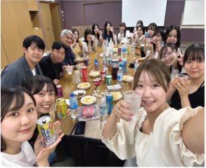 野末ゼミの学生と教員（中央奥の男性）が、一つの長いテーブルを囲んで集まり、懇親会のような雰囲気で笑顔でカメラにポーズしている集合写真（自撮り）。テーブルの上には、たくさんの飲み物（缶、ペットボトル）やスナック菓子が並べられ、皆が飲み物を持っている様子が写っている。