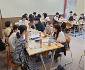 学生たちが、明るい教室でテーブルを囲んで座り、話している様子の写真。手前のテーブルには、資料や飲み物、紙皿などが置かれており、リラックスした雰囲気で会話に集中している。奥にも複数の学生のグループが見える。