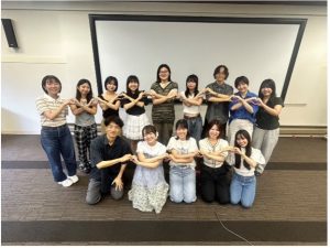 伊藤先生と13名が2列に並んで横の人とハートを作っている写真