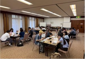 大学のゼミの活動中写真。広い会議室のような場所で、多くの学生がテーブルを囲み、資料を見ながらグループで話し合っている様子。