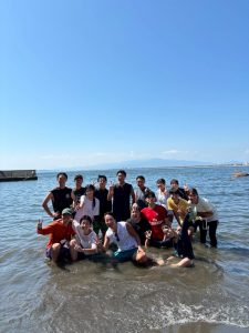 岡田ゼミの海での集合写真。青い海と空の下、Tシャツなどを着た学生たちが楽しそうに水に浸かっている様子。