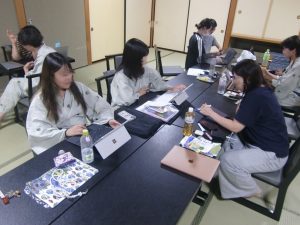和室で、浴衣（または作務衣）姿のゼミ生たちが長いテーブルを囲み、ノートパソコンやタブレット端末を使って作業（またはディスカッション）を行っている、合宿中の様子。