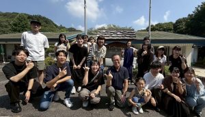 明るい日差しの下で、学生、教員、そして小さな子どもを含む様々な世代のメンバーが、宿泊施設の前で親しげにポーズをとっている記念写真。