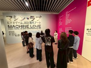 美術館にて、展覧会「MACHINE LOVE ビデオゲーム、AIと現代アート」の導入部分で、大勢のゼミ生が解説を聞いている様子の写真。
