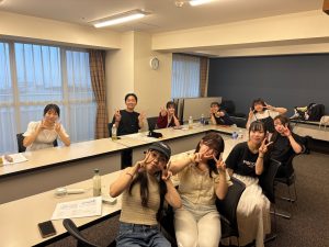 コの字に並べられた長机のある部屋で、9人のゼミ生たちが机を囲んで座り、笑顔でピースしている写真。