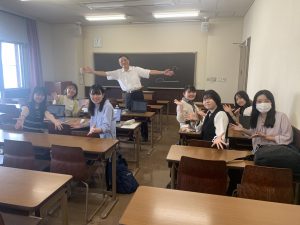 教室で撮影された、中村先生と学生7名の集合写真。中央奥の黒板の前で先生が両腕を広げ、学生たちは机に座ったまま、手を広げたりピースサインをしたりして楽しそうにポーズをとっている。