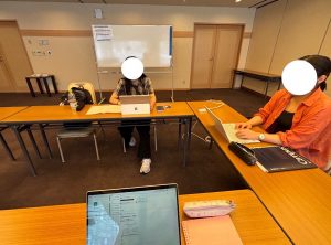 会議室で、2人の人物が離れた机に座り、パソコンを使った作業やミーティングを行っている風景。手前には別のパソコンの画面が見え、奥にはホワイトボードが設置されている。顔は白丸で隠されている。