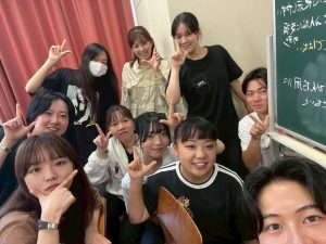 杉岡ゼミの集合写真1。教室の隅に集まり、自撮りで杉岡先生と9人のゼミ生がそれぞれ手でLのポーズをとっている。