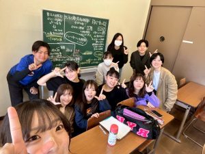 杉岡ゼミの集合写真2。教室の後方で、杉岡先生と10人のゼミ生がそれぞれ手でLのポーズをとっている。後ろの黒板にはたくさんの文字や丸、矢印が書かれている。