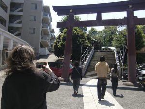 白金氷川神社と思われる大きな朱色の鳥居をくぐり、階段を上ろうとする4人の写真。手前にはカメラを構える人物（撮影者）の背中。左奥にはマンションなどの現代的な建物が見えている。