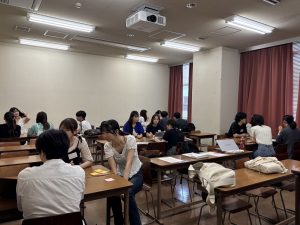 大学の教室で、大勢のゼミの学生たちが机を向かい合わせにして、グループディスカッションやワークショップを行っている様子。