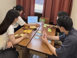 教育工学・教育方法ゼミの少人数グループワークの様子。4人の学生が机を囲み、ノートパソコンのプレゼンテーション画面を参照しながら、付箋（ポストイット）を使ってアイデア出しを行っている。