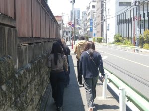 晴れた日の街路を歩く、美術教育ゼミ生4人の後ろ姿の写真。右側には車道と建物、左側には石垣と木塀が続いている。
