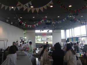横浜の象の鼻テラスの建物の広いイベントスペースで、大勢の来場者がアートマーケットや展示を見ている様子。奥の大きな窓からは屋外の緑が見える。